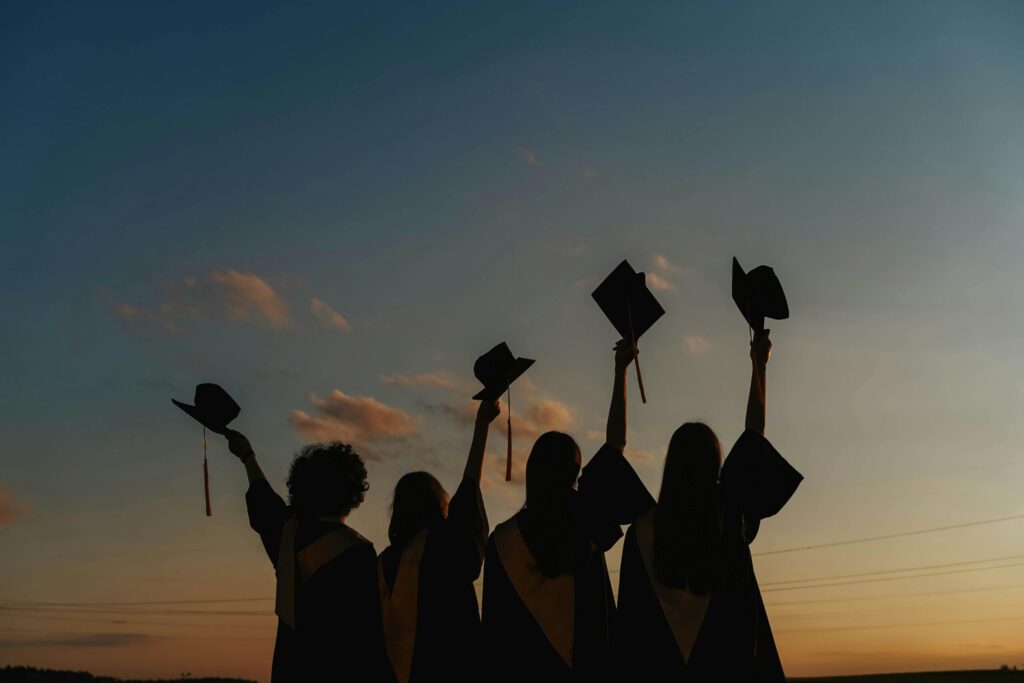 four college graduates in graduate gowns in front of a sunset, throwing their hands in the air